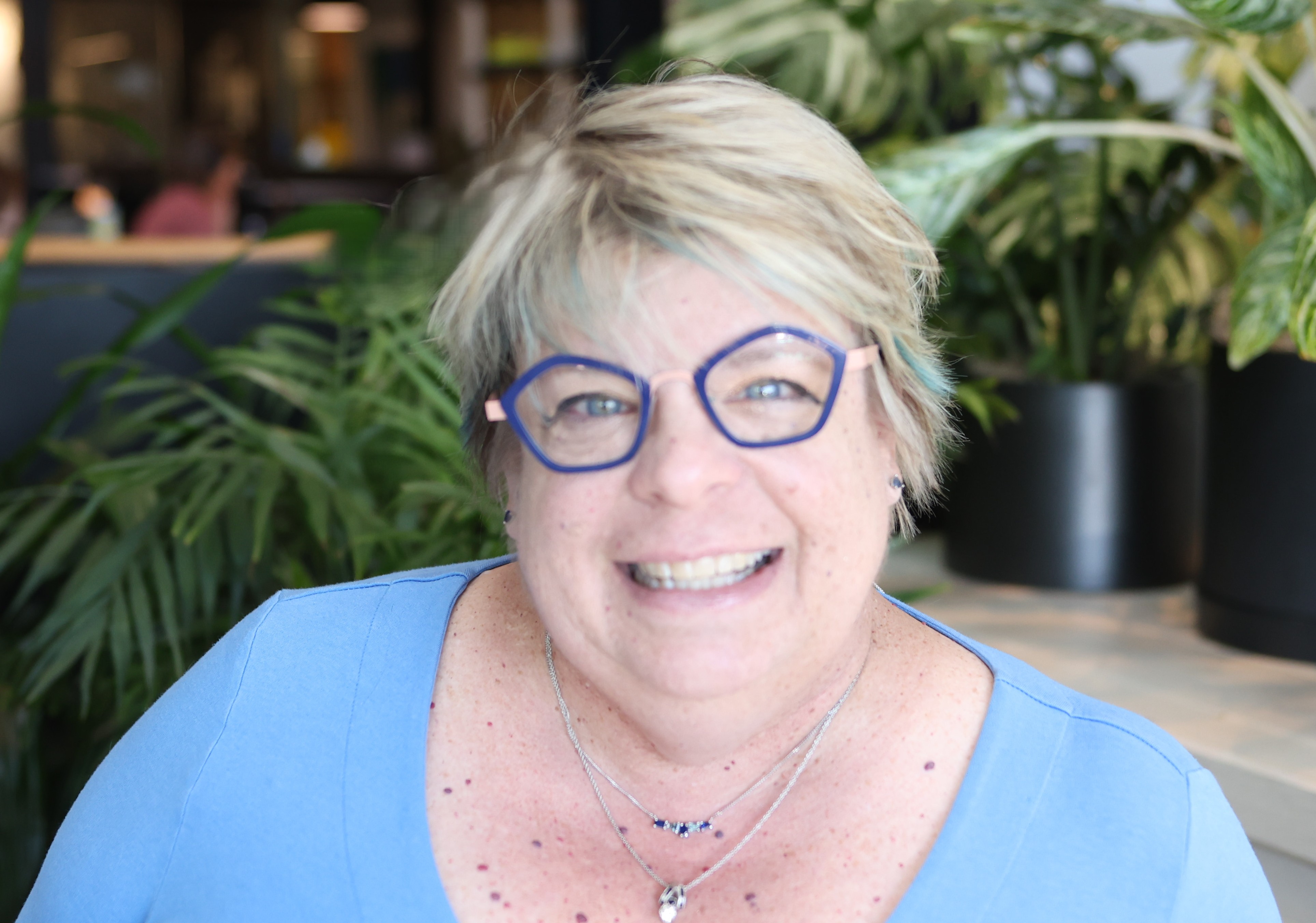 Smiling middle-aged woman with short blonde hair and glasses wearing a blue top and two necklaces in a plant-filled indoor setting.