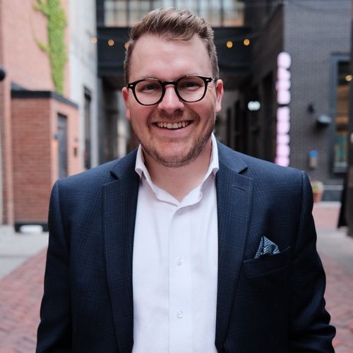 Derek Simancas in suit and glasses in metropolitan area smiling for the camera.