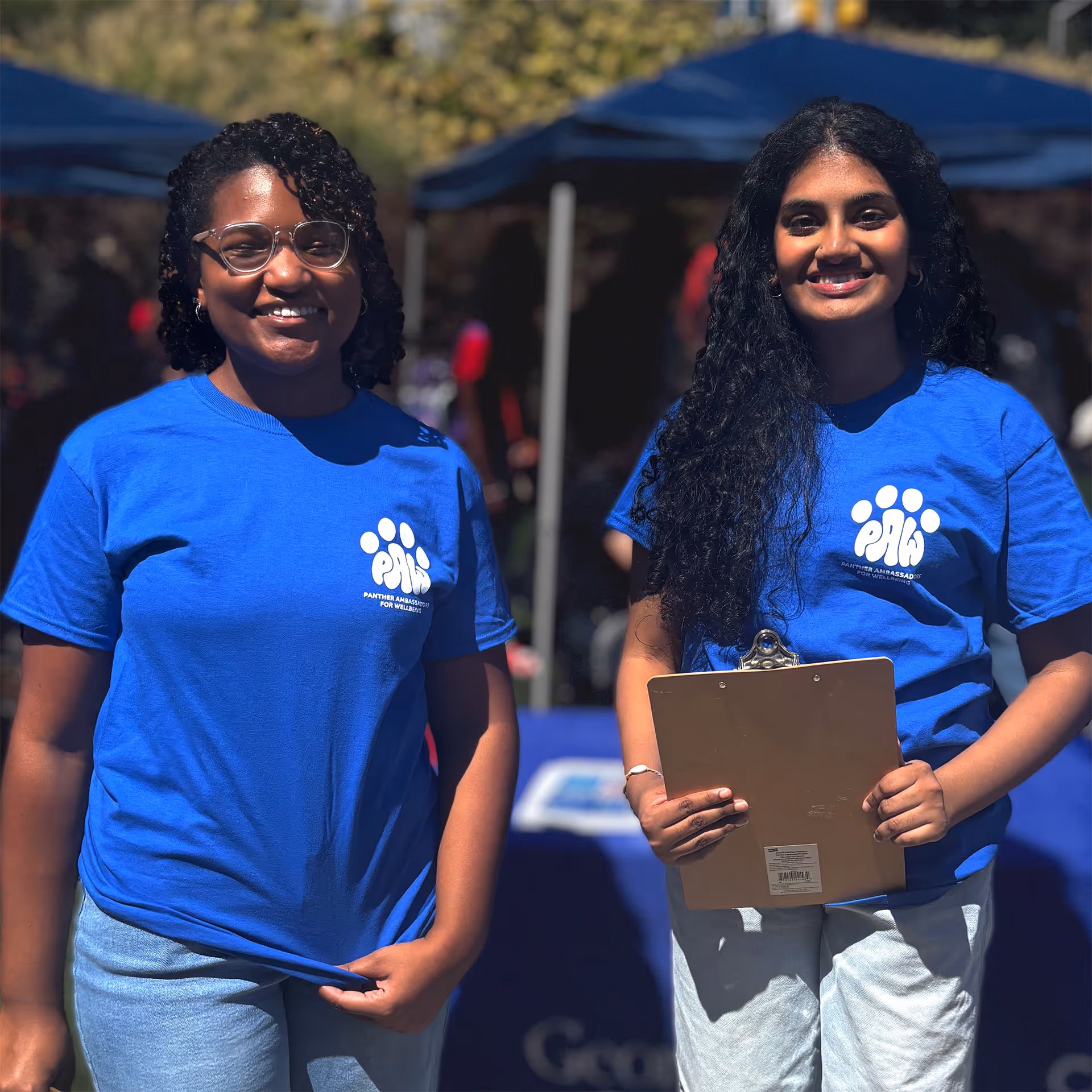 Two women smiling wearing blue PAWS shirts, one holding a clipboard at an outdoor event.