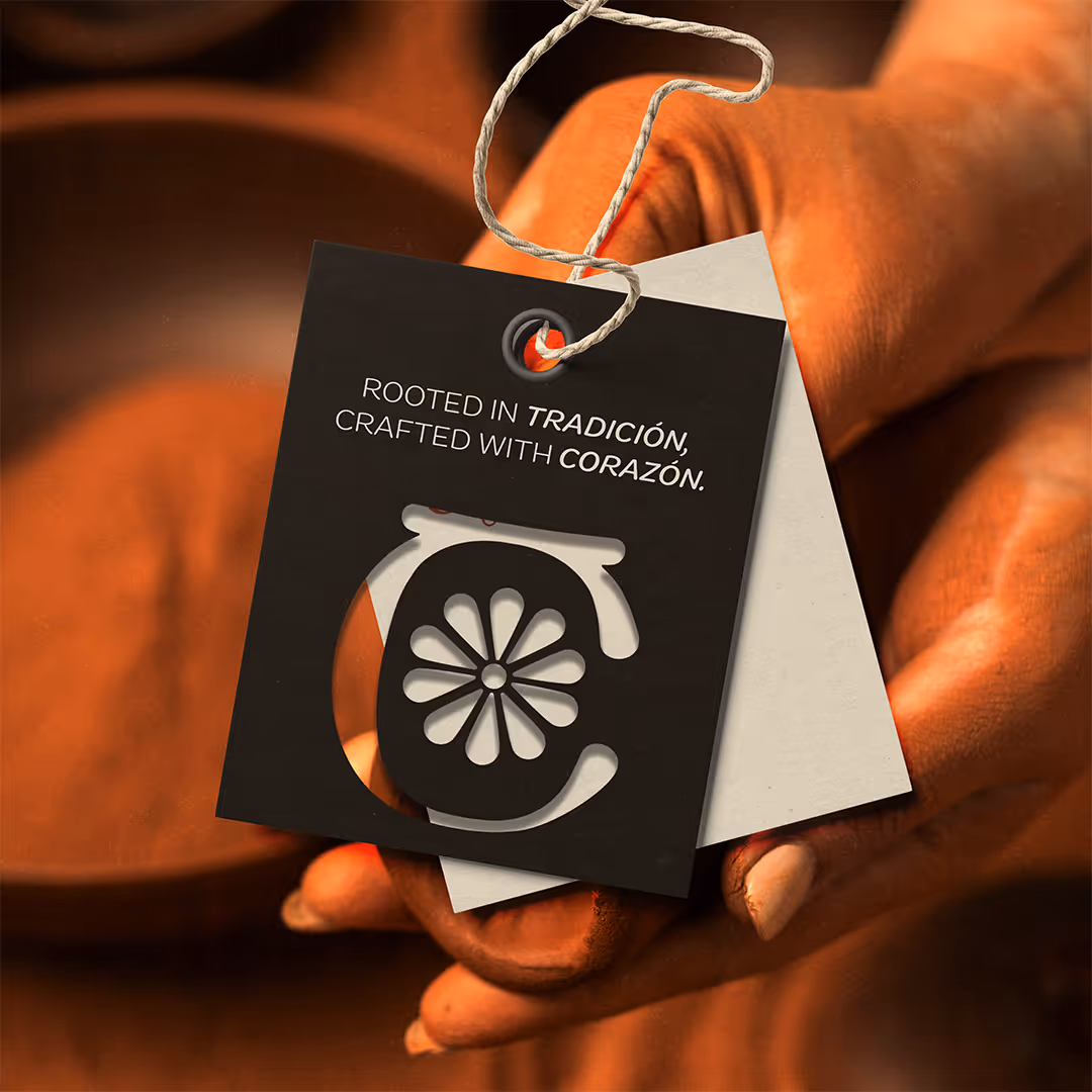 Hand holding a tag reading: Rooted in tradición, crafted with corazón, with decorative cutout design shaped like a clay pot.