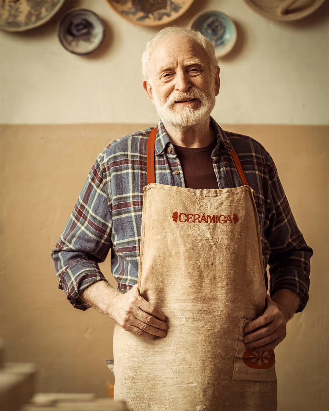 Smiling elderly man in a plaid shirt wearing a ceramiga apron in a pottery studio.