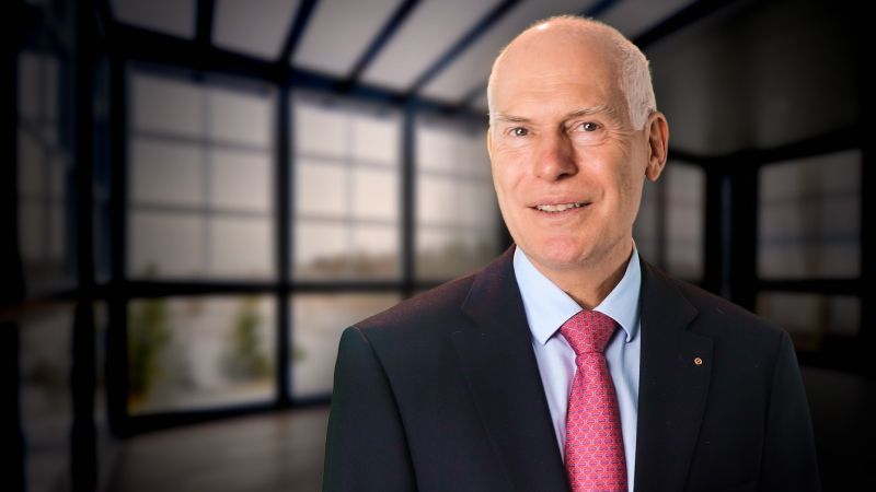 Porträt eines älteren Mannes mit grauem Haar, der einen schwarzen Anzug, ein blaues Hemd und eine rote Krawatte trägt, vor einem modernen Büro mit großen Fenstern.