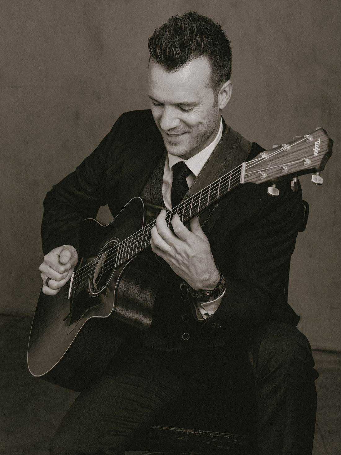 Dylan Galvin smiling while playing acoustic guitar in a three-piece suit, black and white portrait.