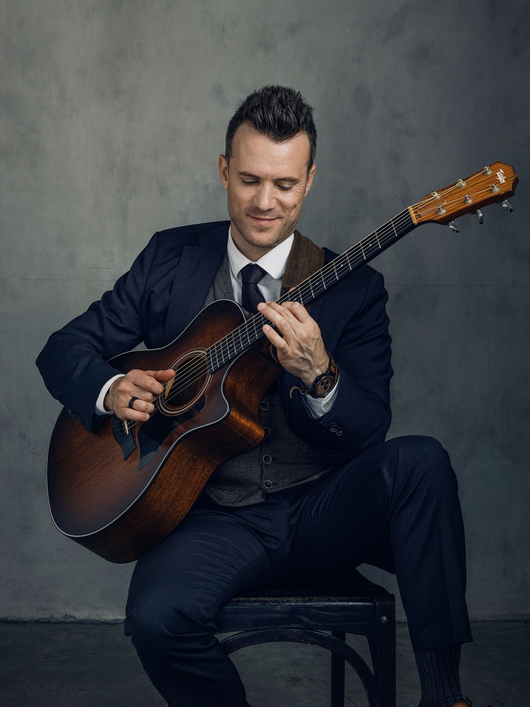 Dylan Galvin, solo acoustic guitarist, seated in a navy suit and grey waistcoat, playing guitar against a concrete wall.