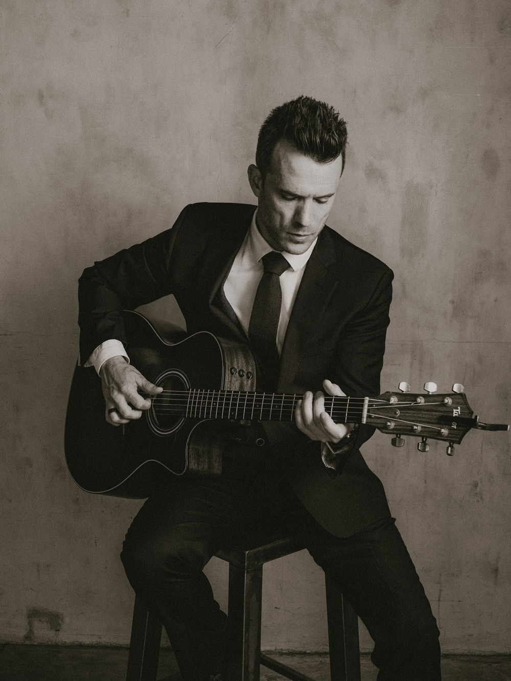Dylan Galvin playing acoustic guitar in a dark suit and tie, seated, head lowered over the instrument.