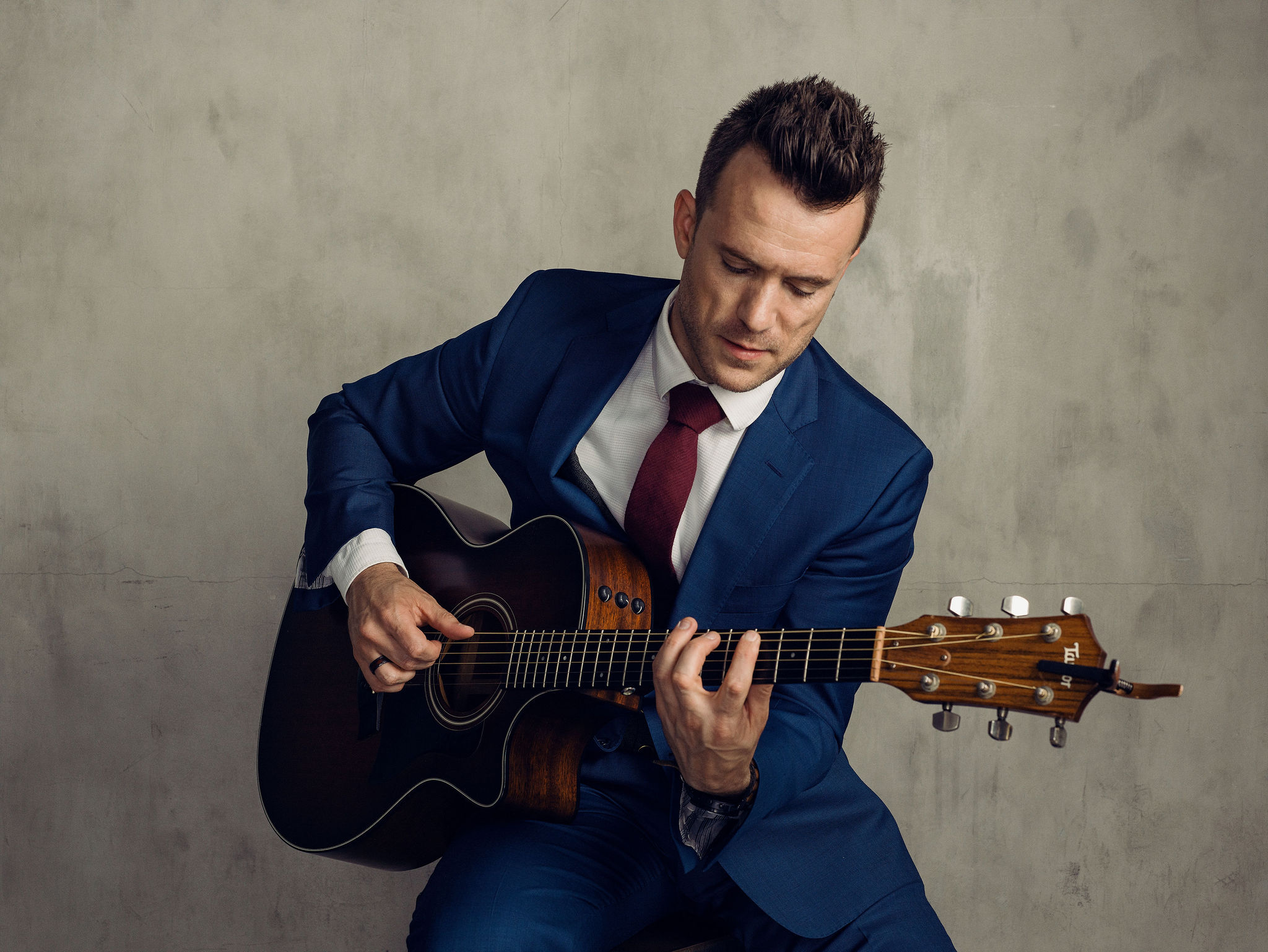 Studio portrait of Dylan Galvin performing acoustic guitar against a concrete wall backdrop