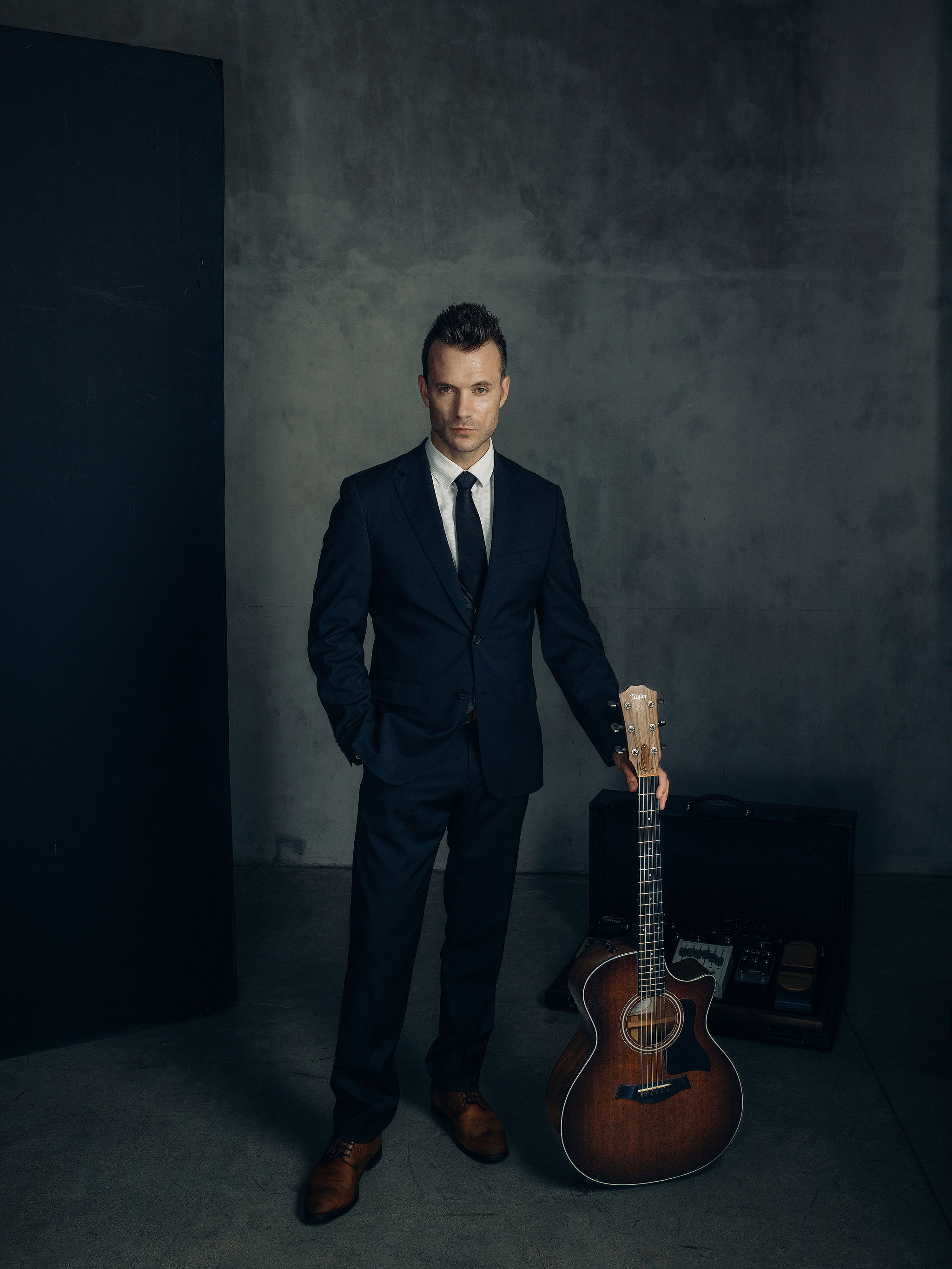 A solo performer in a suit posing with an acoustic guitar case in a moody studio portrait, representing the compact solo or hybrid format next to a full band's stage plot
