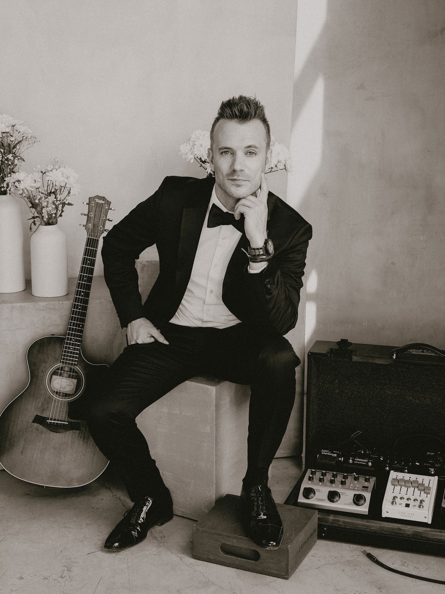 A guitarist in a tuxedo and bow tie sits for a black-and-white studio portrait, acoustic guitar and amp beside him.