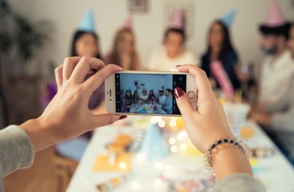 Hands holding a phone to photograph friends in party hats around a birthday table