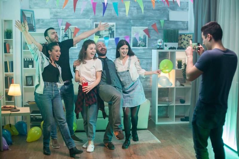 Friends laughing and posing at a birthday party while a man takes their photo with a phone