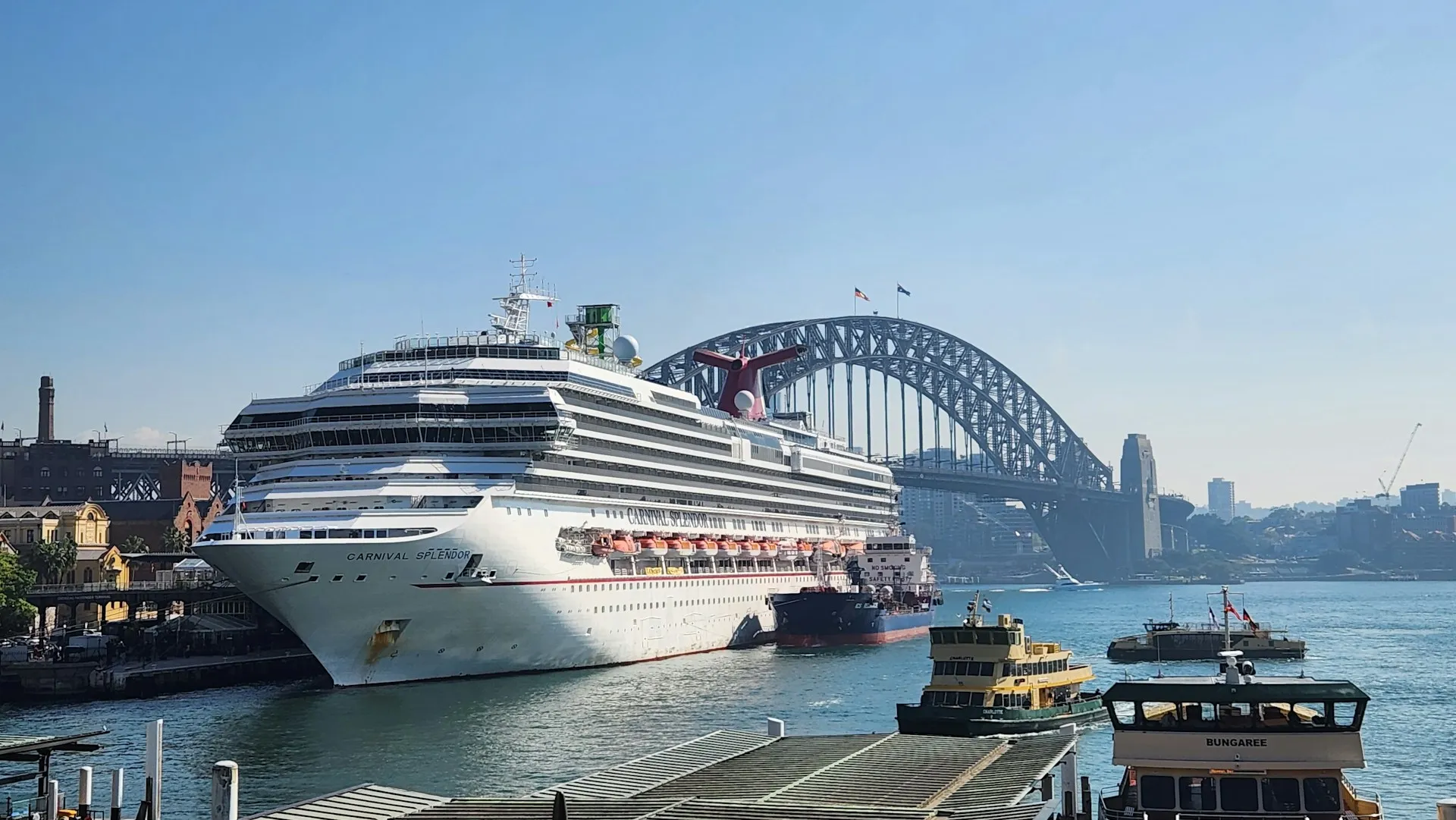 Cruise ship Carnival Splendor docked at the Sydney Overseas Passenger Terminal in Circular Quay, with the Sydney Harbour Bridge in the background and ferries in the foreground.