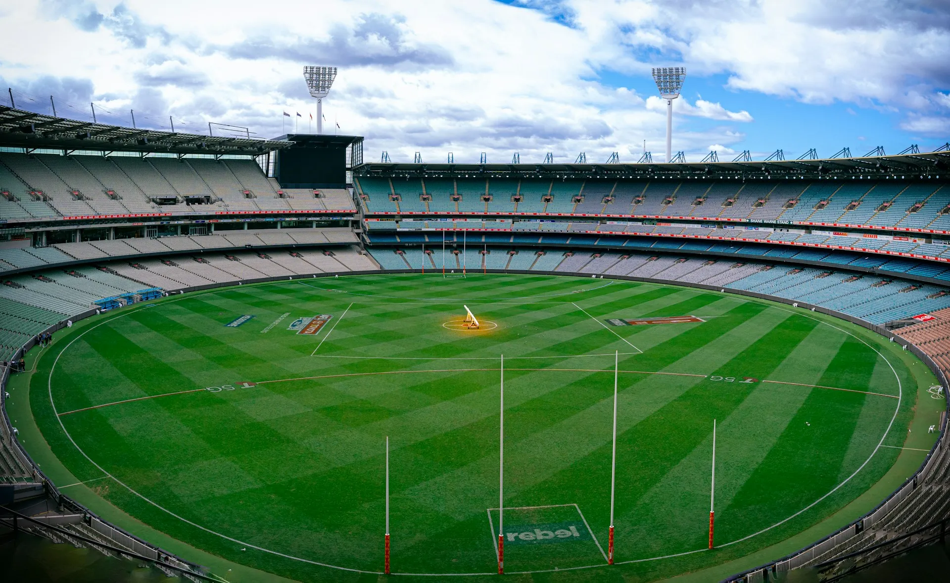 Wide-angle view of an empty Melbourne Cricket Ground (MCG) with neatly mowed green grass and AFL goalposts under a cloudy sky.