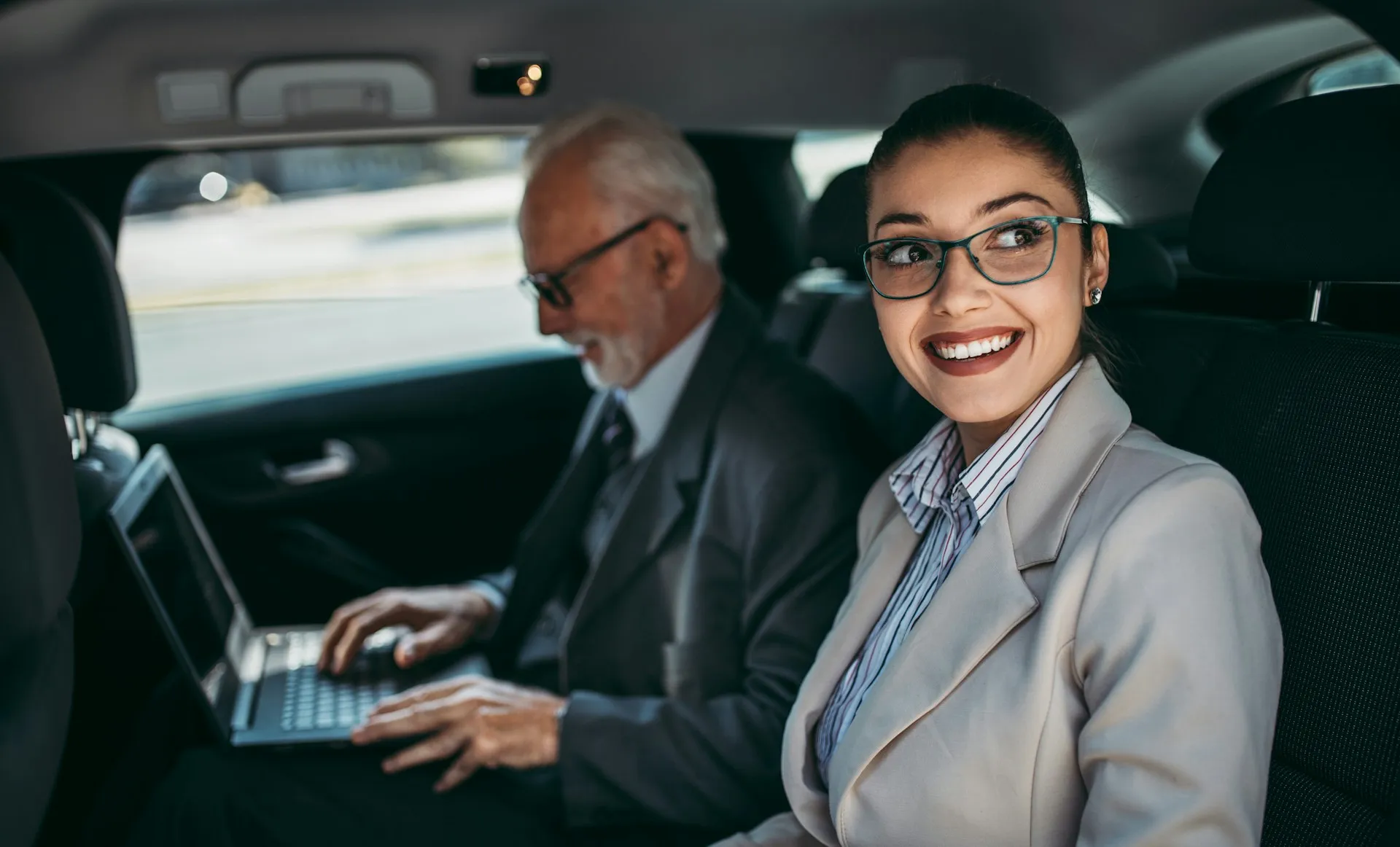 Two business professionals sitting comfortably in the back of a chauffeured vehicle, one smiling and relaxed while the other works on a laptop, illustrating seamless group transport coordination.