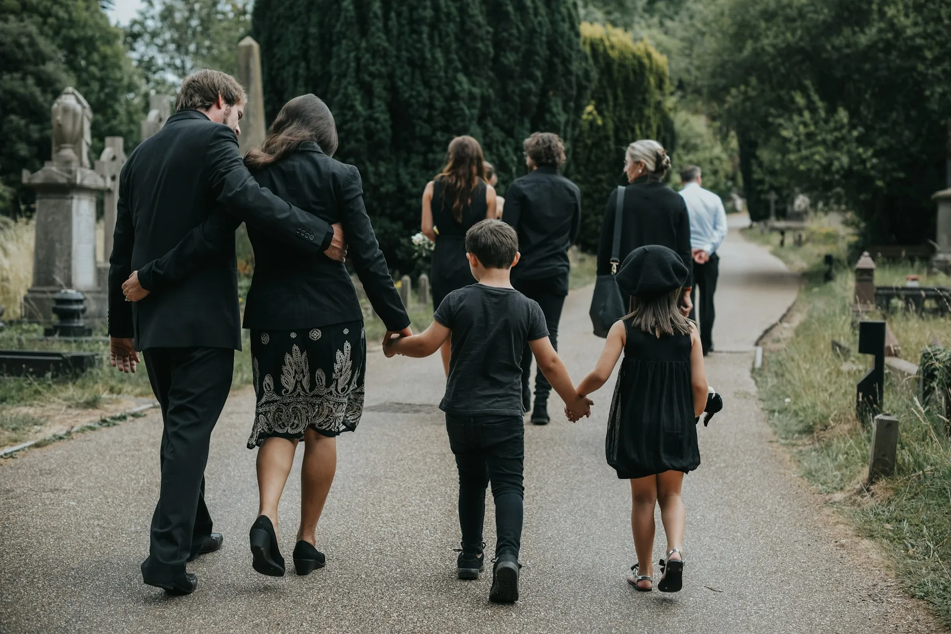 Family members, including young children, walk hand-in-hand through a cemetery in formal attire, symbolising togetherness and support during a funeral — reflecting the importance of private, respectful family transport.