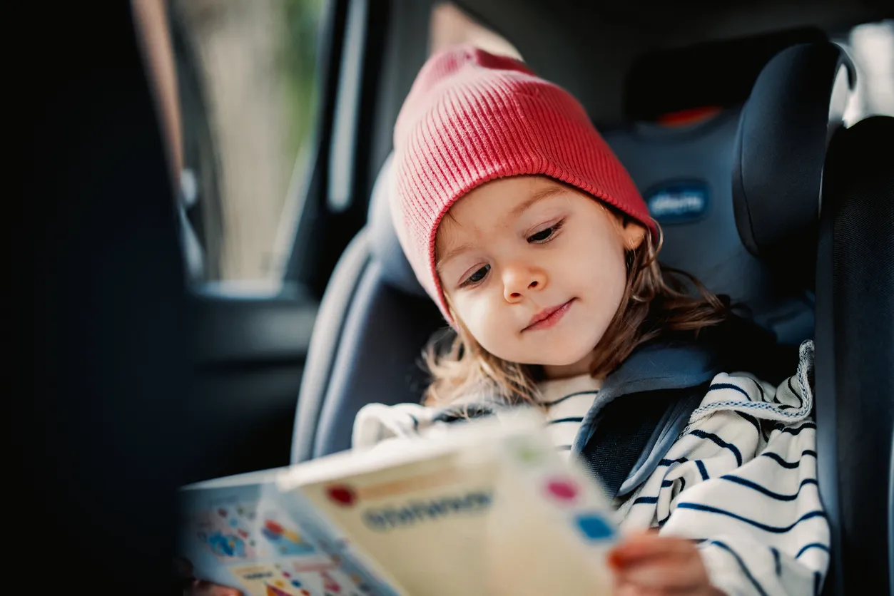 Young girl safely seated in a car seat reading a book, representing comfort, safety, and peace of mind at the heart of Evoke’s family transport services.
