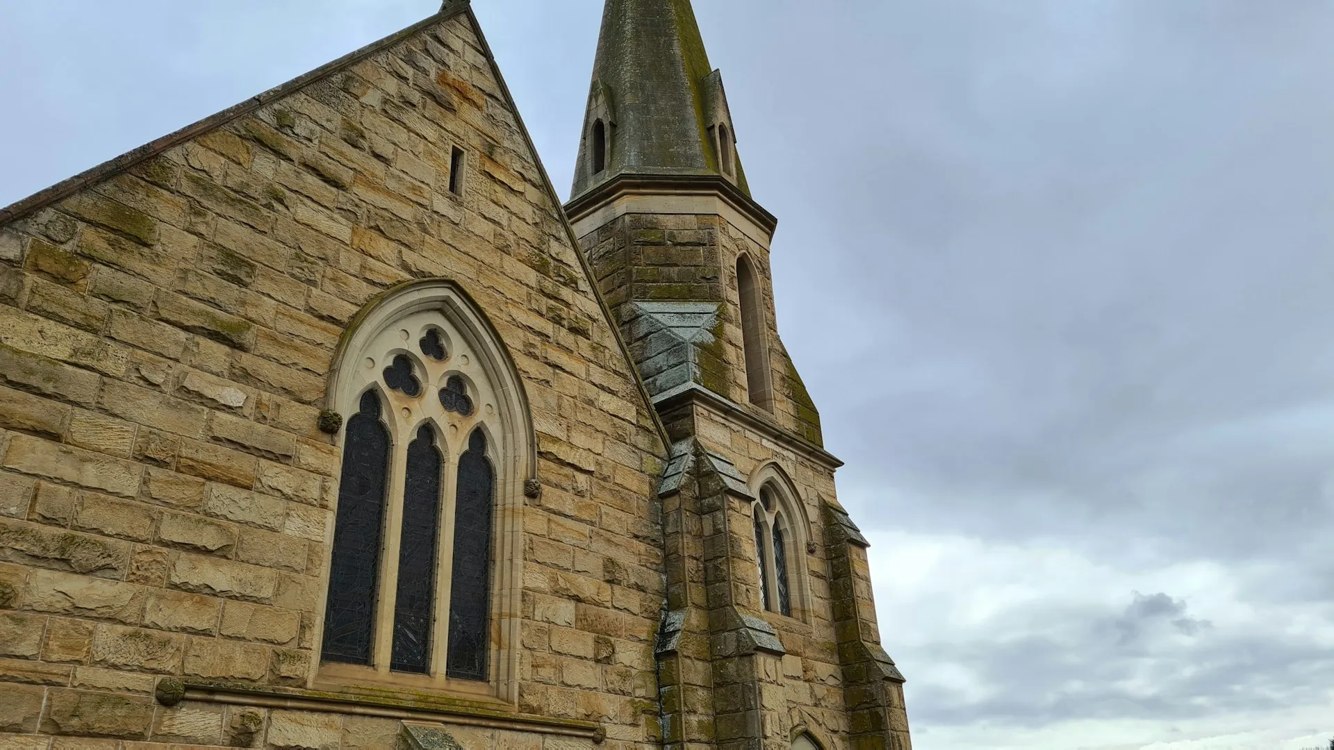 Sandstone church with gothic-style arched windows and a tall spire, set against an overcast sky.