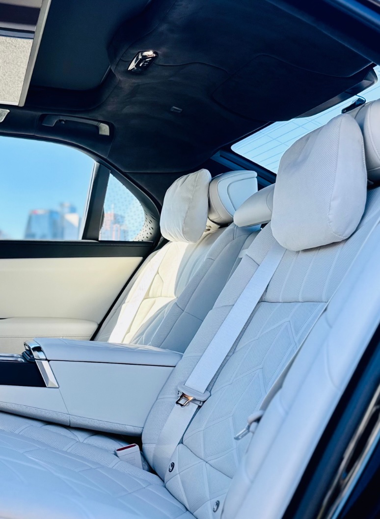 Rear interior view of the BMW i7 showcasing white quilted leather seats, a centre armrest, and headrest cushions, with city buildings visible through the window.
