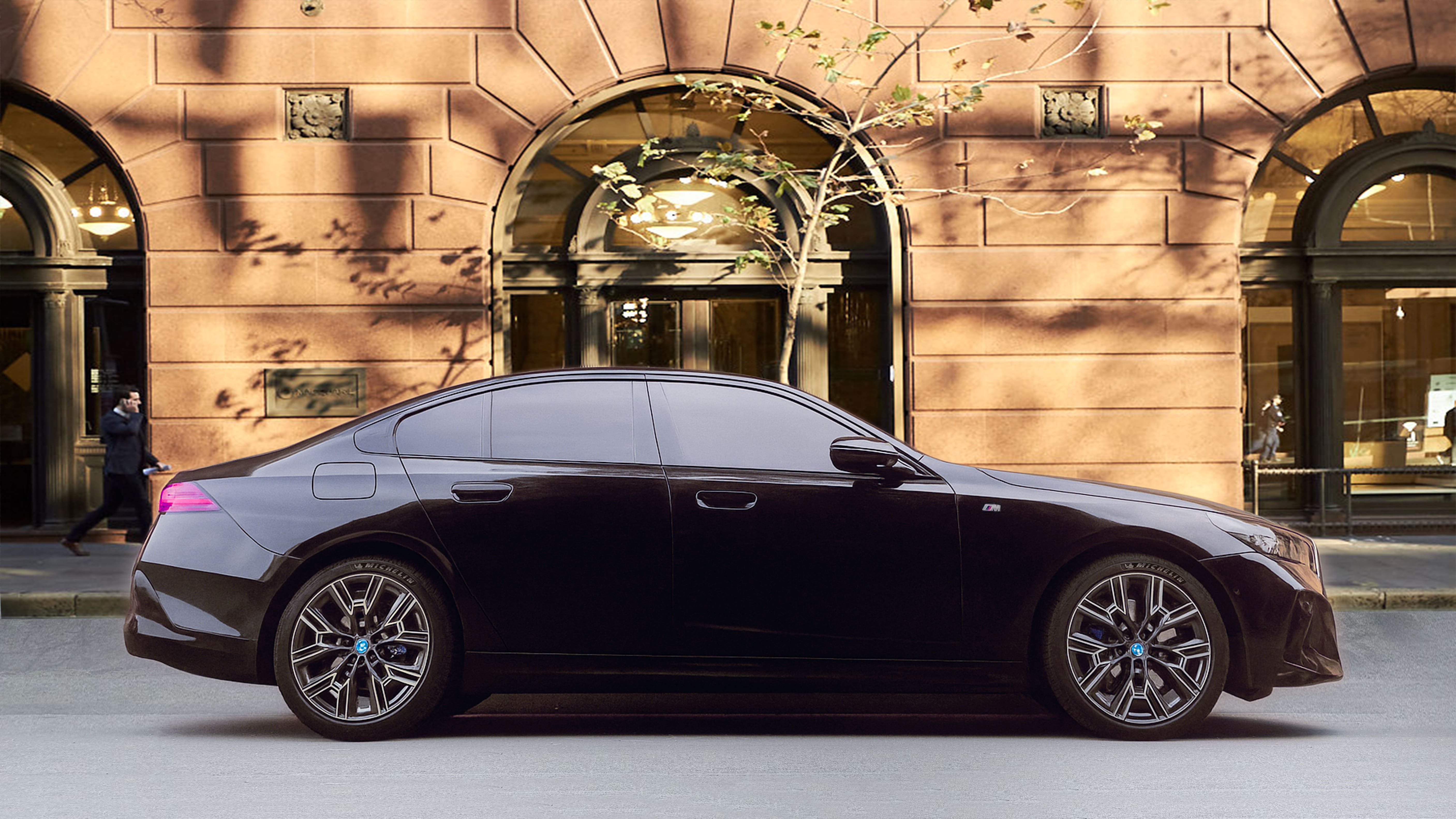 Side profile of a black BMW i5 parked on a city street in front of a sandstone building with arched windows.