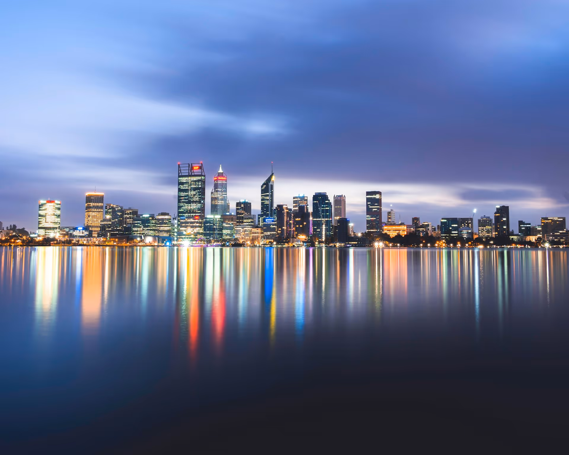 Perth CBD city skyline at dusk with illuminated buildings reflecting in the calm waters of the Swan River.