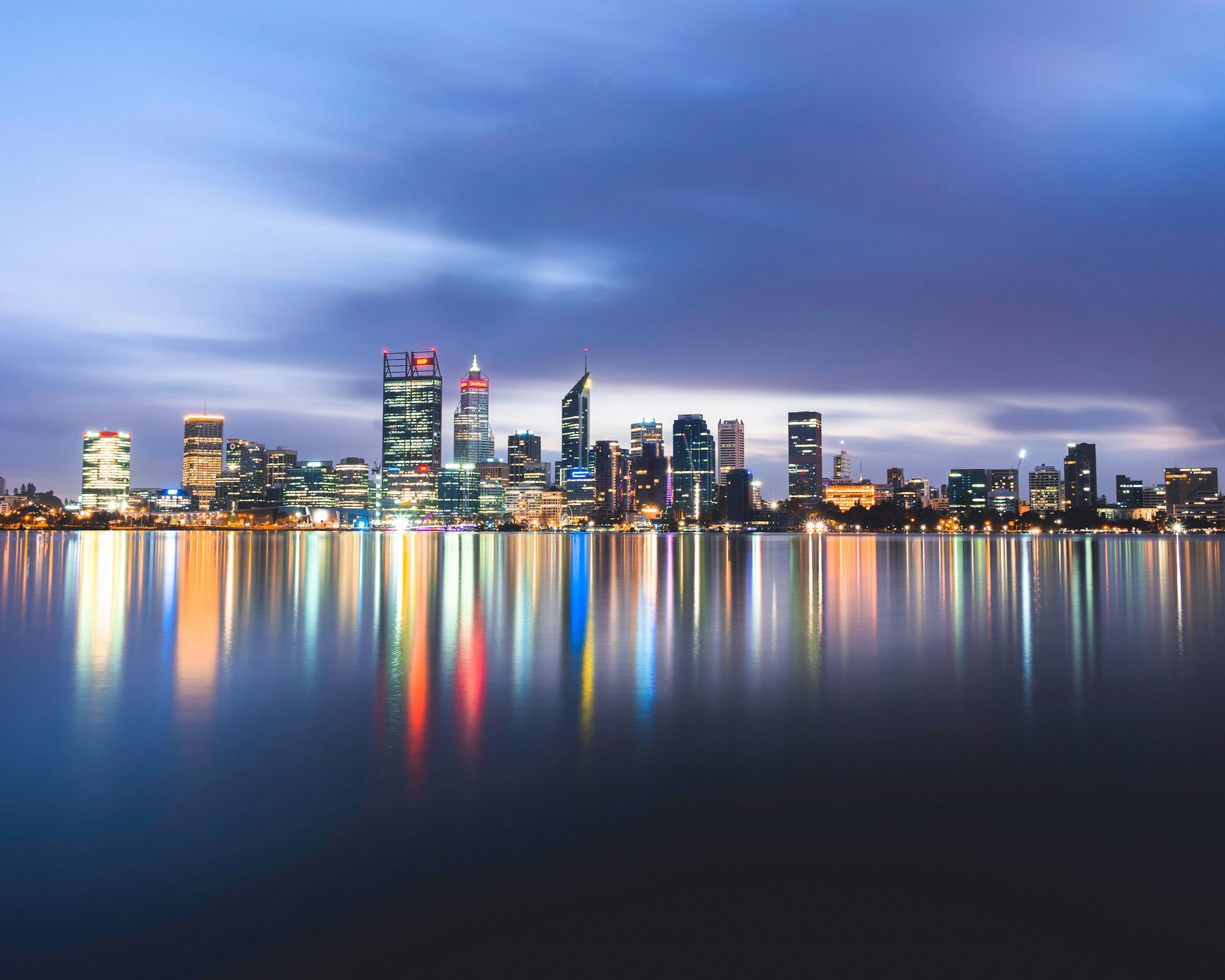 Perth CBD city skyline at dusk with illuminated buildings reflecting in the calm waters of the Swan River.