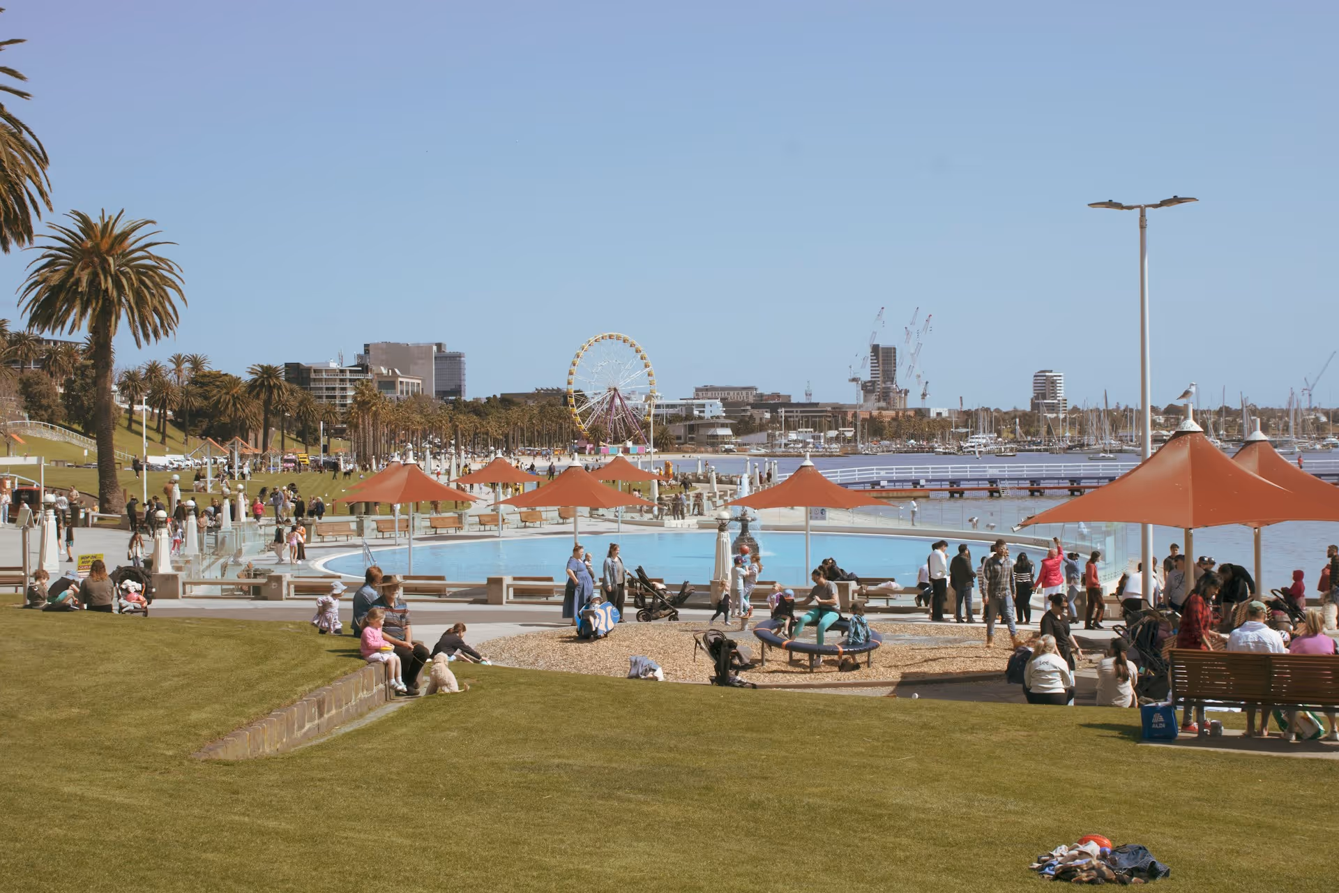 Crowds enjoying the waterfront, pools, and grassy areas at Eastern Beach Reserve in Geelong, Victoria, with the ferris wheel in the background.