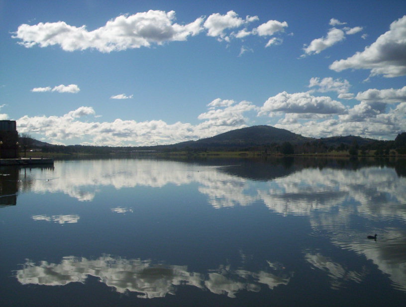 Lake Tuggeranong in Canberra with still water reflecting clouds and surrounding hills, captured on a clear day. Credit: Cliffzart, Wikimedia Commons: https://commons.wikimedia.org/wiki/File:Lake_tuggeranong_reflection.jpg