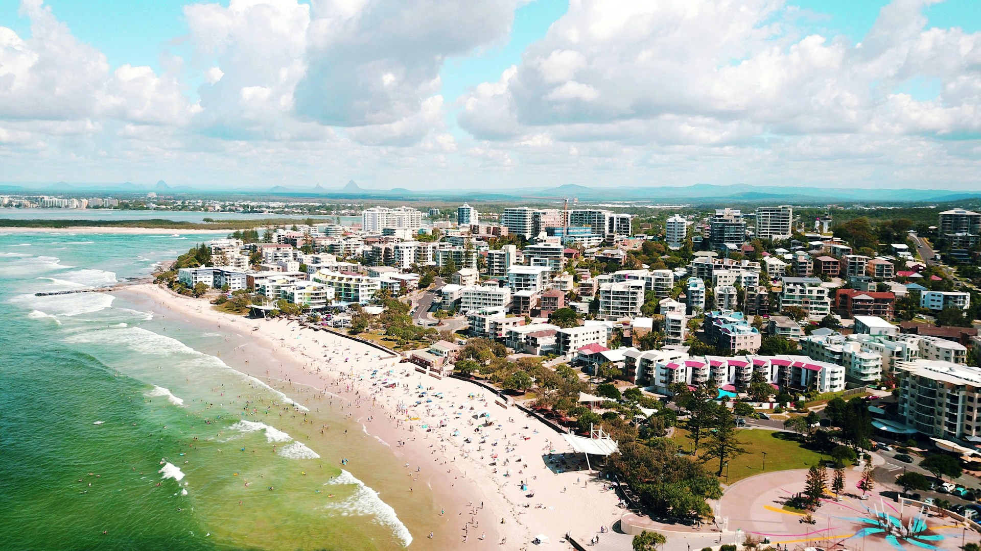 Aerial view of Kings Beach on the Sunshine Coast, Queensland, showing people swimming and relaxing along the sandy shoreline with coastal apartments nearby.