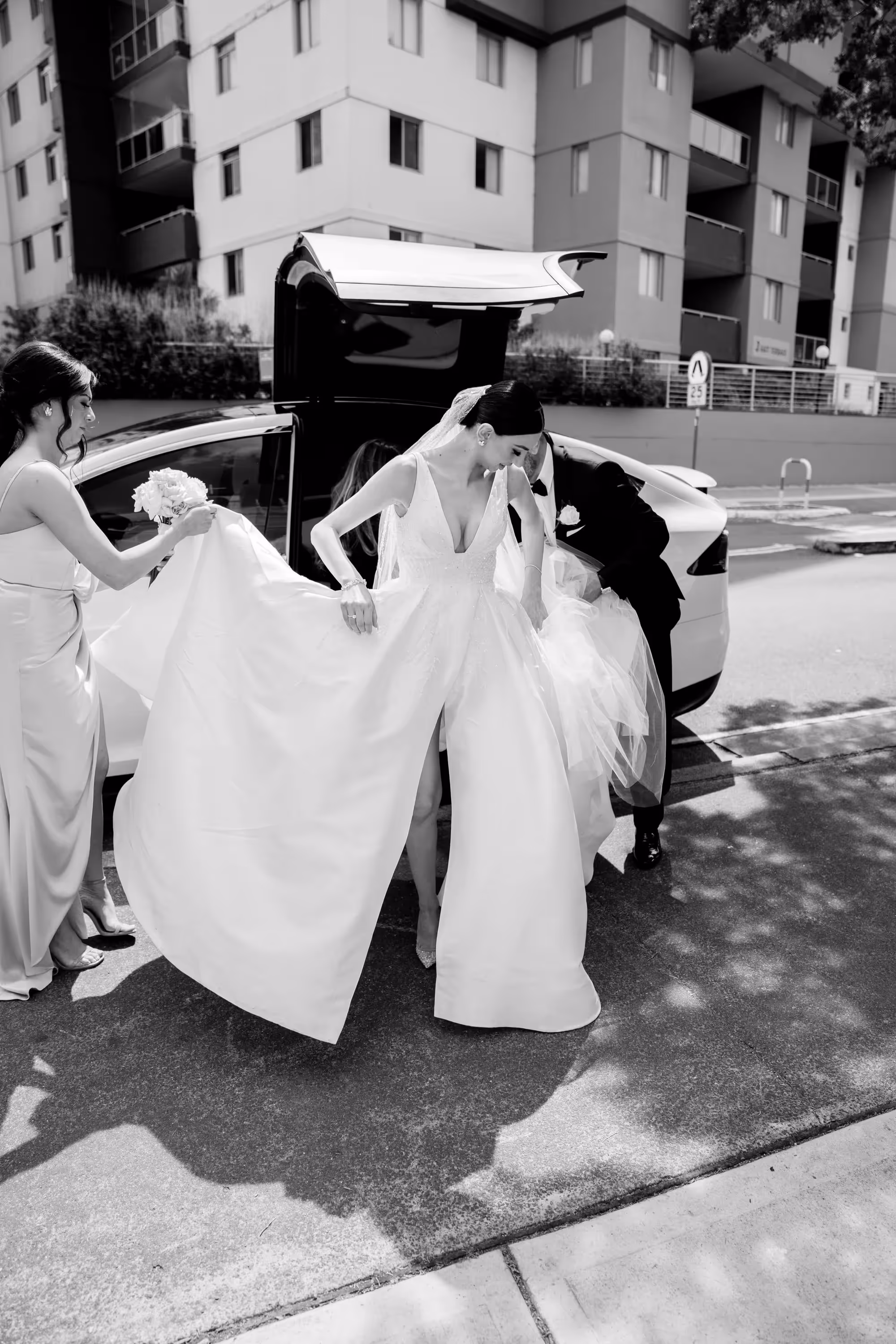 Bride exiting Tesla Model X with falcon wing door open, while being assisted by father and bridesmaid.