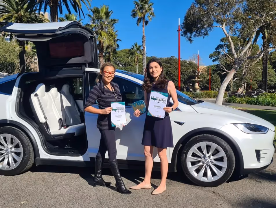 Amanda Choy and Pia Peterson standing next to a white Tesla Model X with falcon-wing doors open, holding awards, highlighting sustainable and eco-friendly recognition.