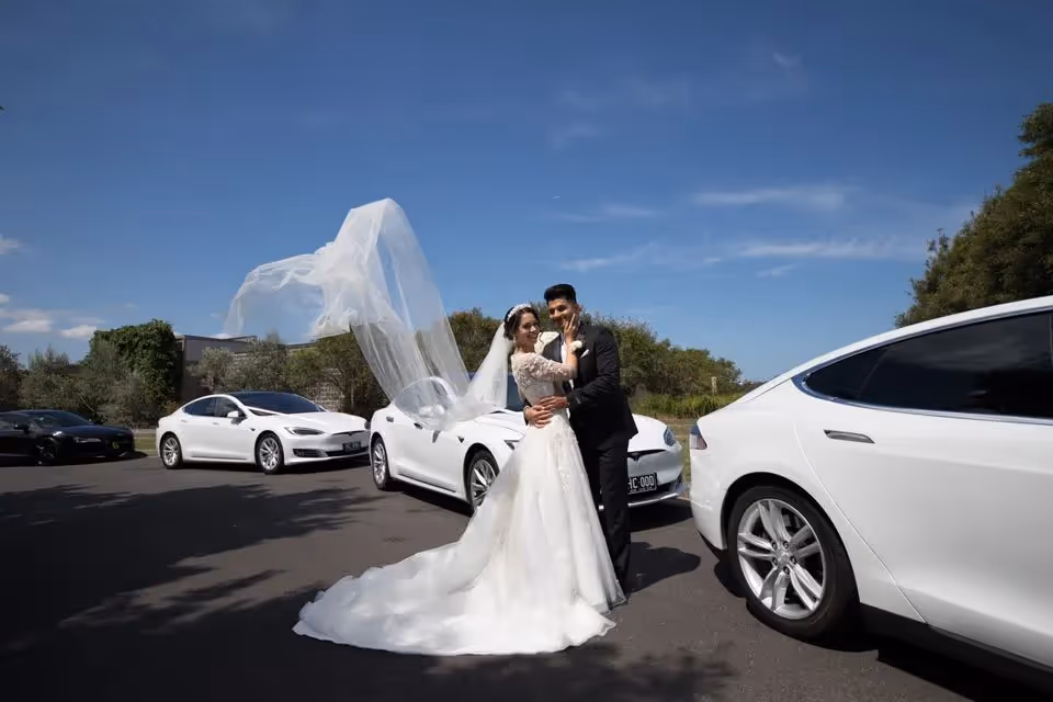 Bride and groom posing in front of white Tesla cars, with the bride's veil blowing in the wind, symbolising a modern and eco-friendly wedding celebration.