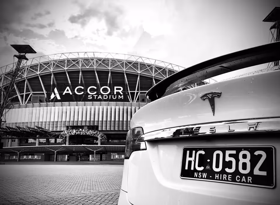 A black and white image showing the rear of a white Tesla Model X with a "NSW - Hire Car" plate parked in front of Accor Stadium.