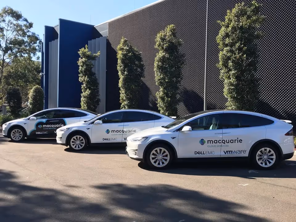 Three white Tesla Model X vehicles branded with "Macquarie Cloud Services" decals, parked in front of a modern building with trees lining the area.