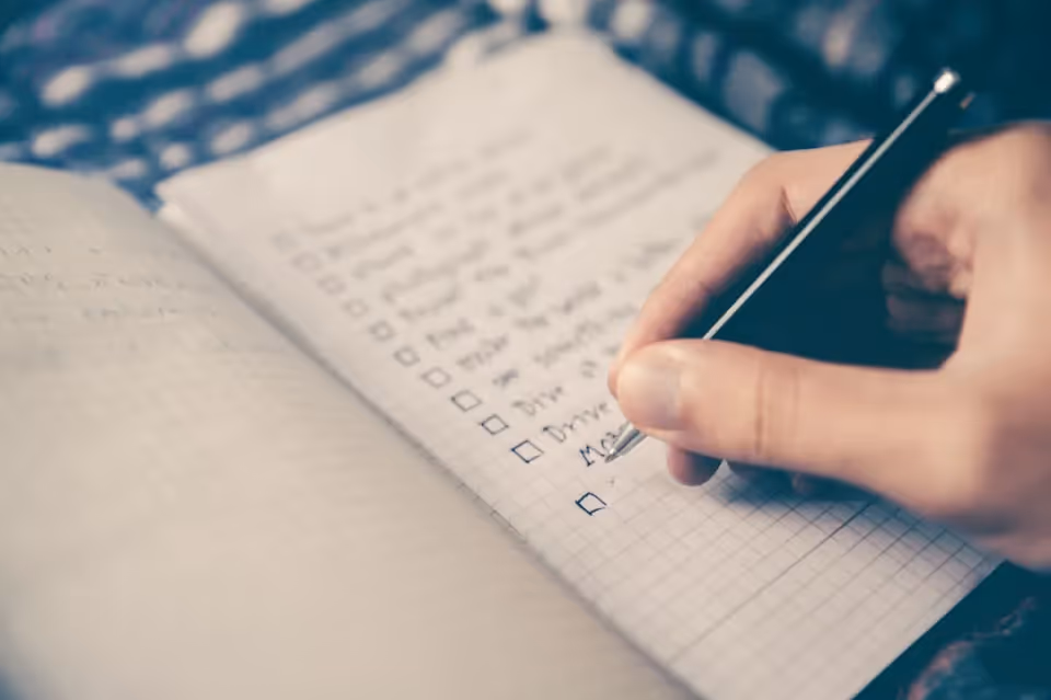 Hand writing a checklist in a grid-patterned notebook with a black pen.