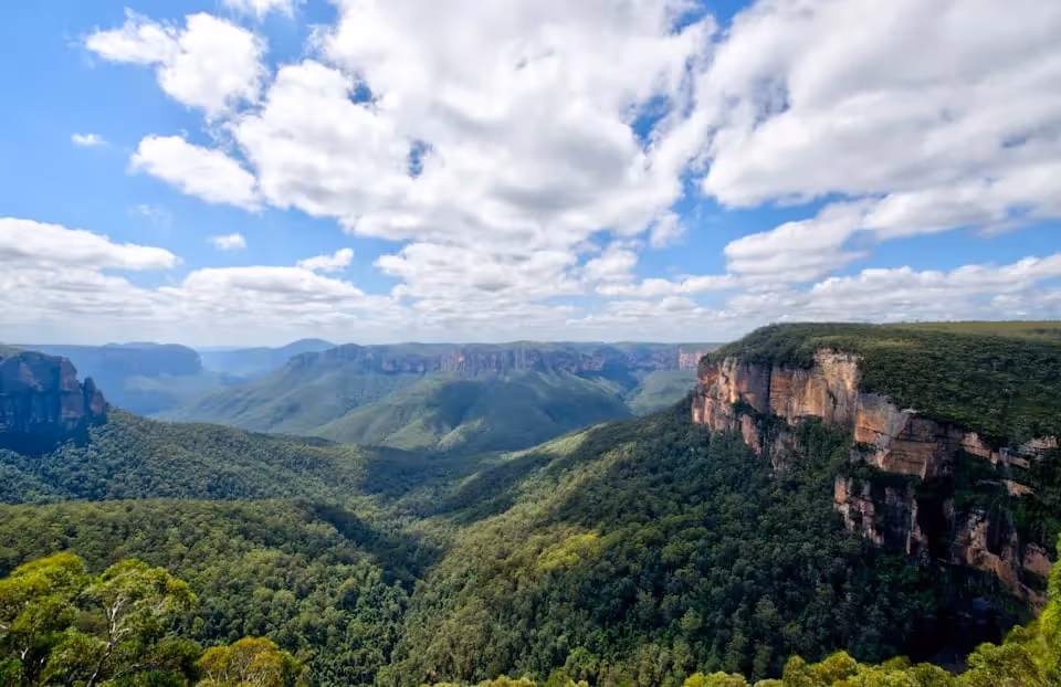 Blue Mountains, Australia, featuring lush green valleys and dramatic sandstone cliffs under a partly cloudy sky.