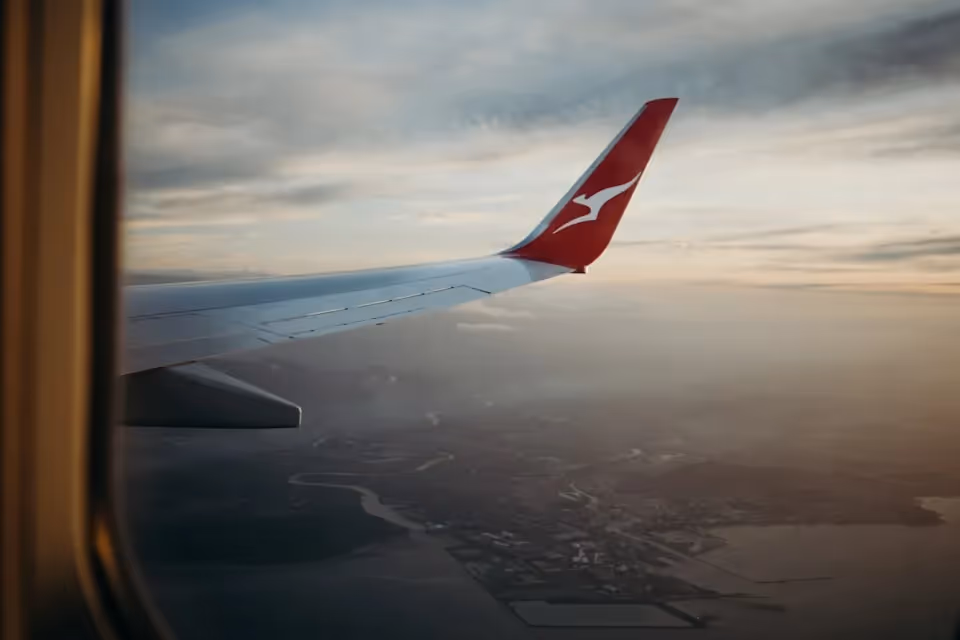 View from an aeroplane window showing a Qantas wing tip flying over a coastal landscape at sunset.