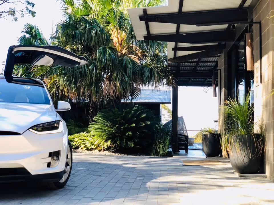 A white Tesla Model X with a falcon-wing door open, parked on a paved driveway at Spicers Sangoma Retreat, with lush greenery and a modern building entrance in the background.