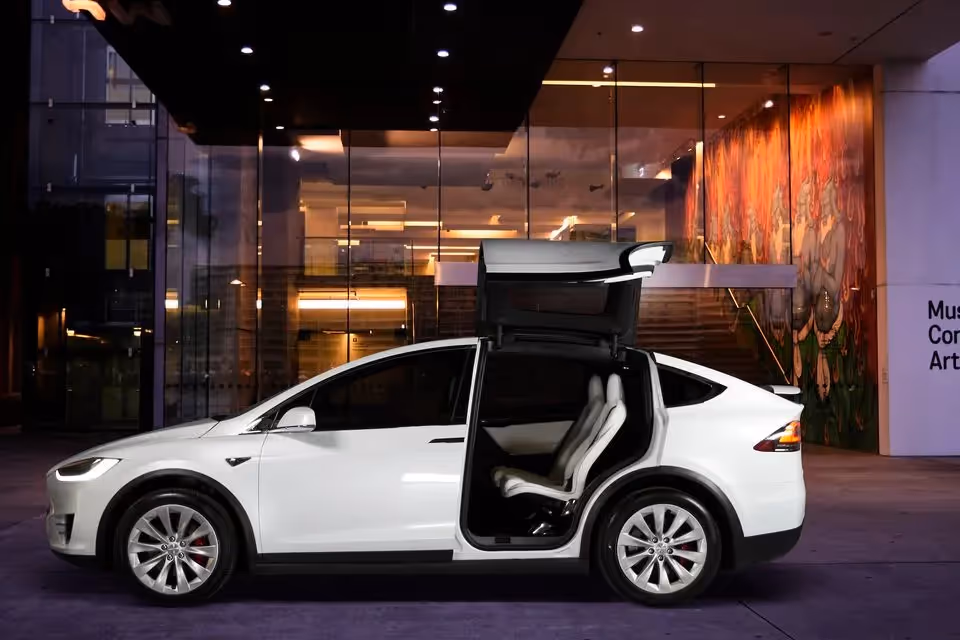 A white Tesla Model X with its falcon-wing door open, parked in front of the Museum of Contemporary Art, which features glass walls and colourful artwork.