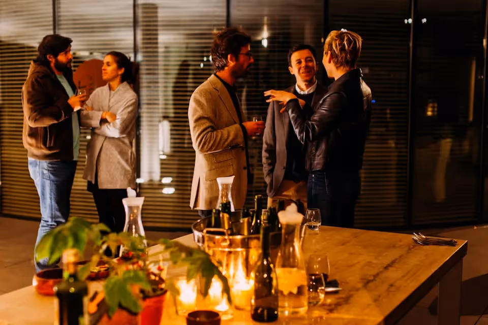 A group of people engaging in lively conversation at an evening social event, with a table in the foreground adorned with bottles, glasses, and lit candles, creating a warm and inviting atmosphere.