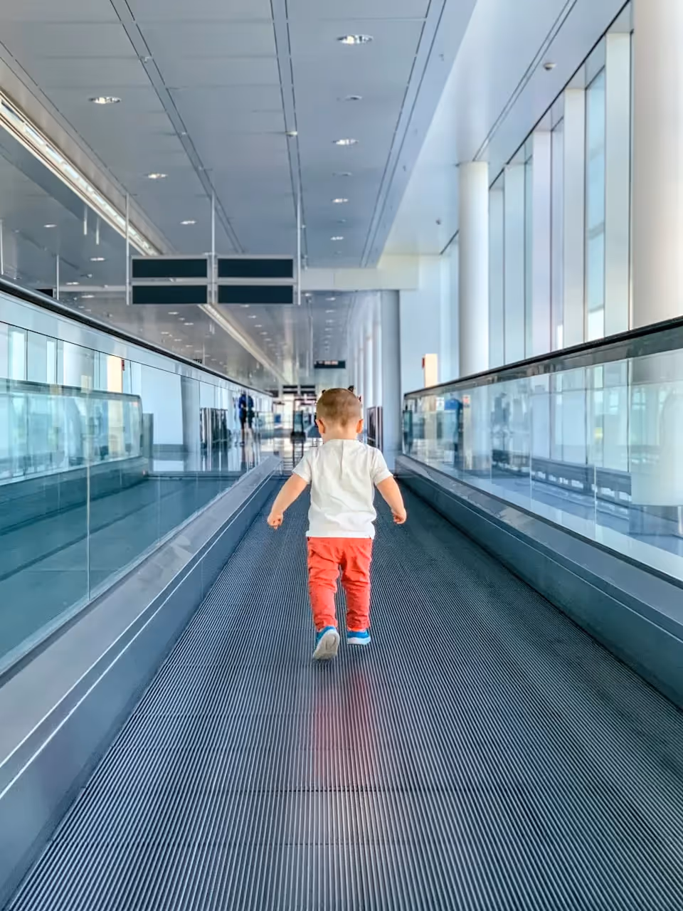 A young child, dressed in a white shirt and red pants, walking on a moving walkway in an empty, modern airport terminal with large windows and a bright interior.
