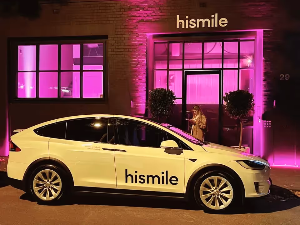 A white Tesla Model X with "hismile" branding parked in front of a building illuminated with vibrant pink lighting, with a woman standing near the entrance.