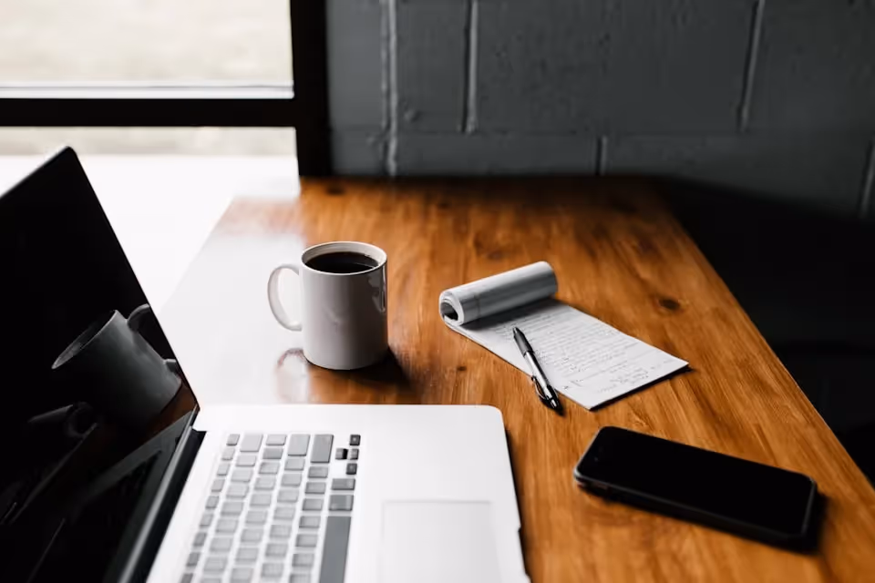 A minimalist workspace featuring an open laptop, a cup of coffee, a notepad with a pen, and a smartphone on a wooden table near a window.
