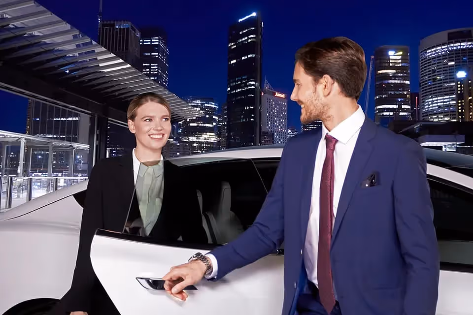 A chauffeur in a suit opening the door of a white car for a smiling businesswoman, with a backdrop of illuminated city skyscrapers at night.