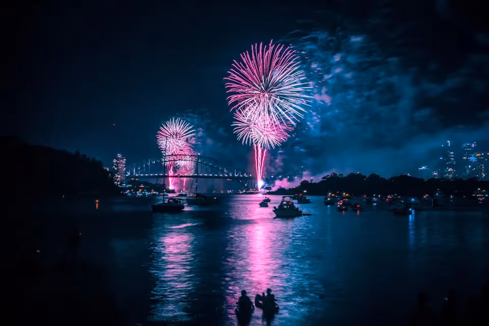 Spectacular fireworks lighting up the night sky over Sydney Harbour, with the Sydney Harbour Bridge and boats on the water reflecting the vibrant pink and purple hues.