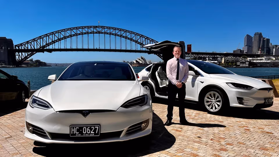 Richard standing in front of two white Tesla cars, one with falcon wing doors open, against the backdrop of the Sydney Harbour Bridge and the Opera House on a sunny day.