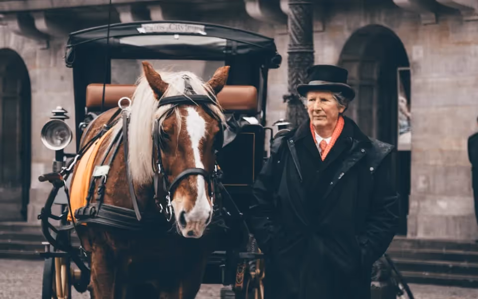 A traditionally dressed coachman standing beside a horse-drawn carriage, with the horse wearing a harness and the carriage set against the backdrop of historic stone buildings.