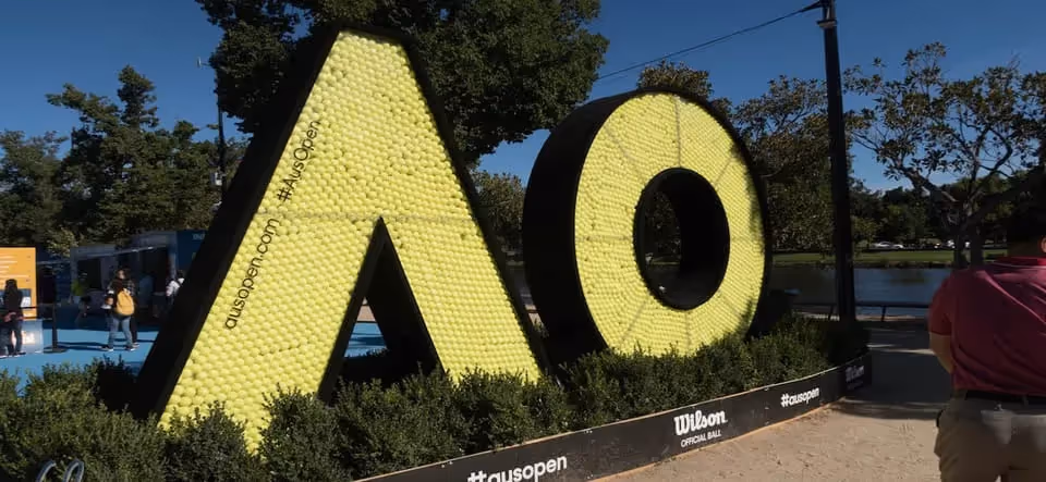 A large sculpture of the letters "AO" made of tennis balls, representing the Australian Open, situated in an outdoor setting with trees and people in the background.