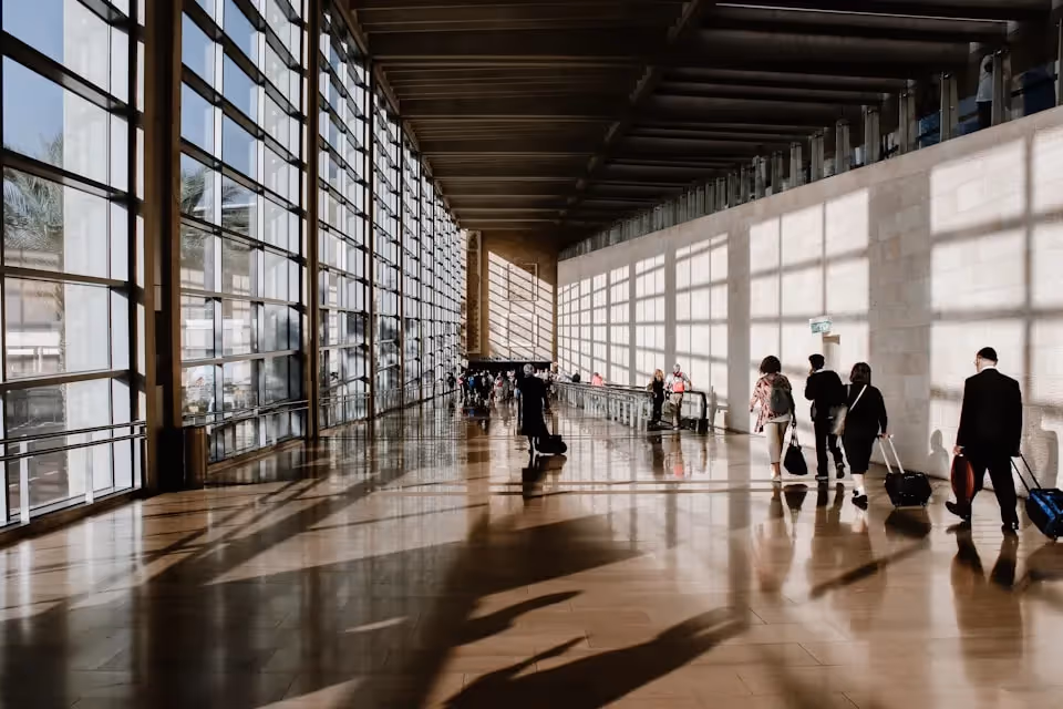 Travellers walking through a spacious, sunlit airport terminal with large windows casting geometric shadows on the polished floor.