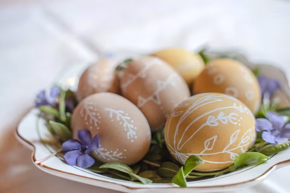 A decorative plate filled with Easter eggs, each adorned with intricate white designs, surrounded by green leaves and purple flowers.