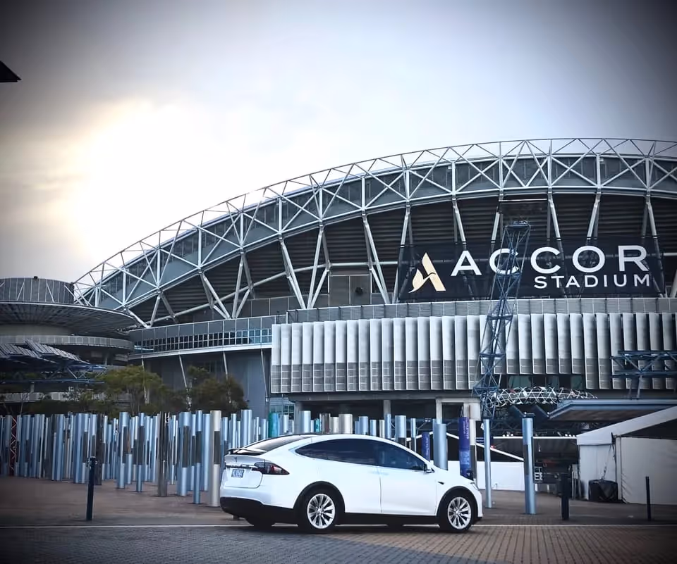 A white Tesla Model X parked in front of the Accor Stadium, with the stadium's distinctive architecture visible in the background under a cloudy sky.
