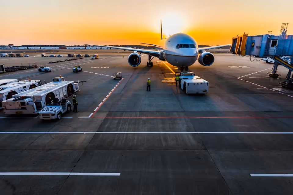 A large commercial aeroplane being prepared for departure at an airport gate during sunset, with ground crew and vehicles in operation, and the sun setting in the background.